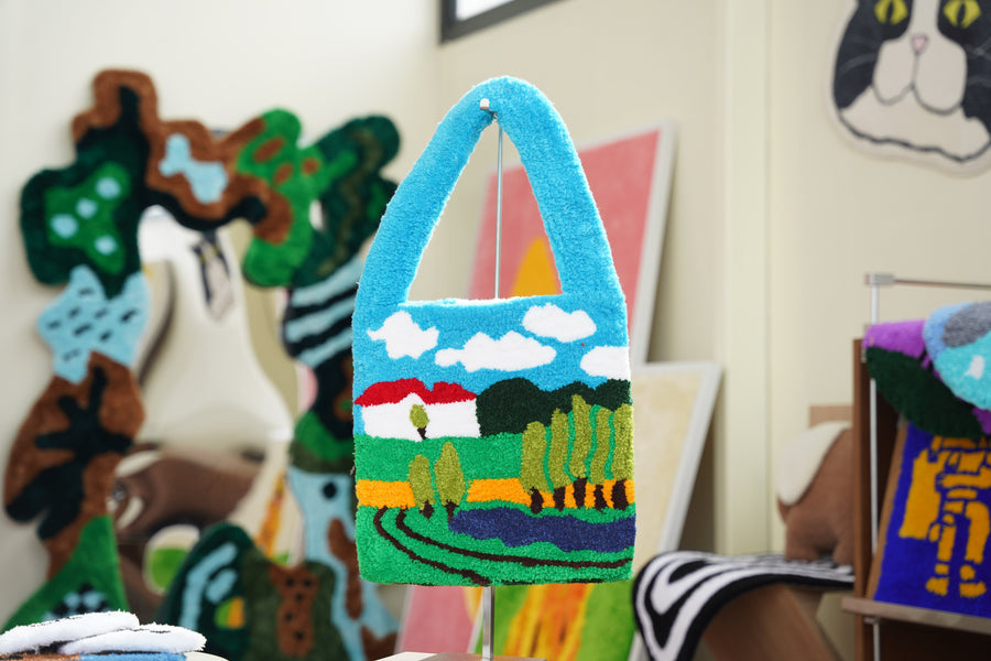 A light blue tufted bag with a countryside scene of a red-roofed house, trees and clouds, hanging in front of colorful tufted rugs in a studio