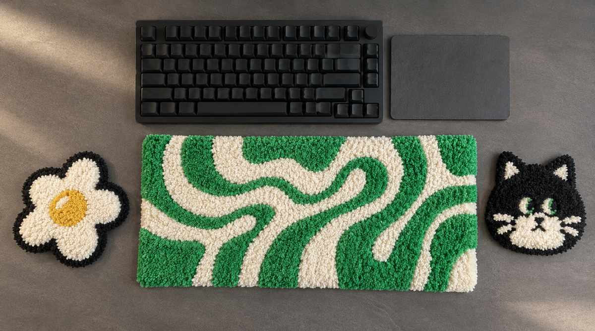 Handmade tufted desk accessories on a dark gray desk: a green and cream wavy wrist rest, a daisy coaster and a black cat coaster, arranged around a black mechanical keyboard and a trackpad